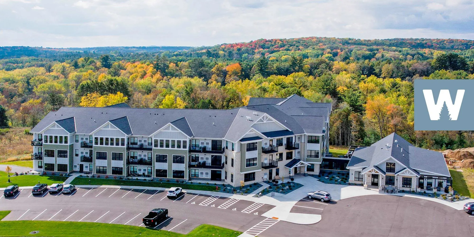 Modern apartment complex with gray roofs along a landscaped lot, autumn-colored forest in the background.