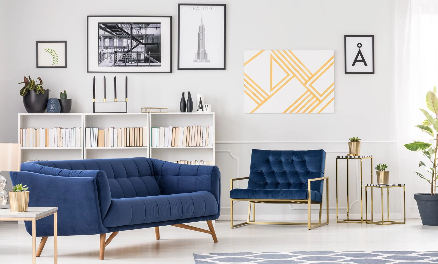 Bright modern living room with a blue velvet sofa and matching armchair, white bookshelves filled with books, and framed wall art in black frames above a light gray wall.